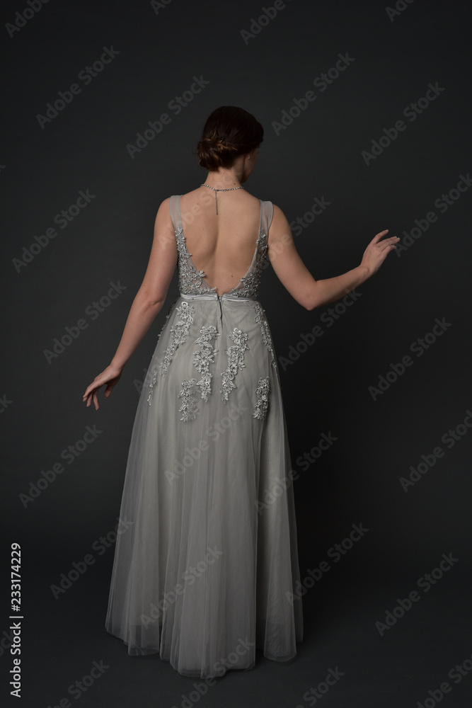 Fototapeta premium full length portrait of brunette girl wearing beautiful long gown, standing pose with background to the camera on grey studio background.
