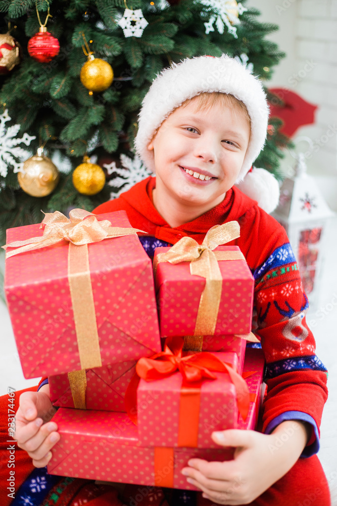 Vertical portrait of cute happy smiling european kid with stack of Christmas presents in red wrapping paper. Boy sits near green decorated holiday tree at home. Color photography. Vertical portrait of cute happy smiling european kid with stack of Christmas presents in red wrapping paper. Boy sits near green decorated holiday tree at home. Color photography.