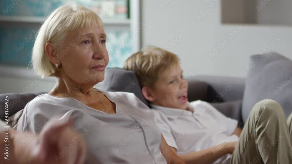 Beautiful senior woman sitting on sofa with little grandson, smiling and watching TV at home Beautiful senior woman sitting on sofa with little grandson, smiling and watching TV at home