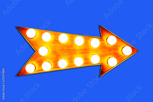 Yellow orange arrow shaped vintage colorful illuminated metallic display direction sign with glowing light bulbs isolated on an intense vivid blue background.