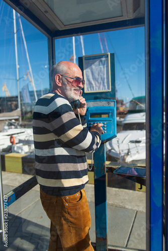 one old man makes a call from a blue payphone booth in a yacht port
