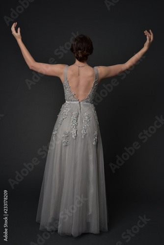 full length portrait of brunette girl wearing beautiful long gown, standing pose with background to the camera on grey studio background.