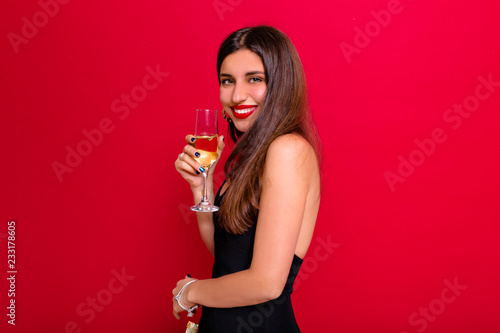 Adorabel lovely woman on red background drinking champagne, celebrating new year, wearing black dress with bare shoulder, happy carnival disco party, sparkling confetti, holding glass, having fun