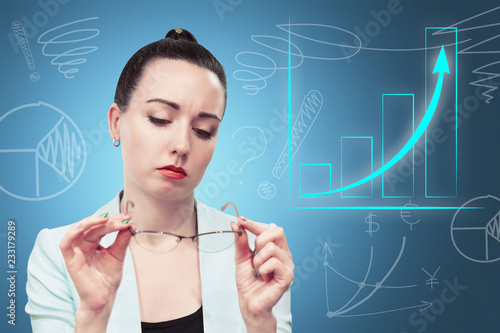 Young black haired business woman in light blue jacket looking at glasses in her hands and virtual business graphs and charts around her against blue background, business concept 