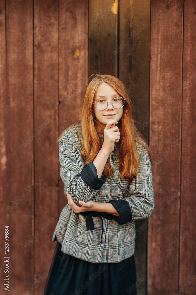 Outdoor portrait of beautiful red-haired preteen girl wearing check coat and eyeglasses, posing next to wooden background Outdoor portrait of beautiful red-haired preteen girl wearing check coat and eyeglasses, posing next to wooden background