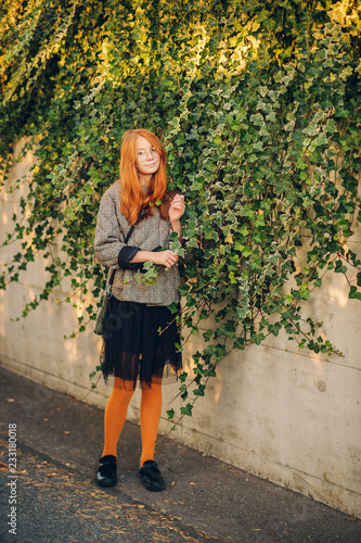 Portrait of adorable little red-haired girl, posing outside, wearing check jacket and black skirt