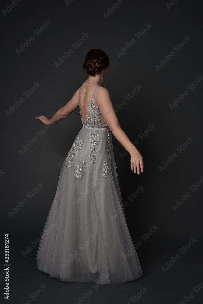 full length portrait of brunette girl wearing beautiful long gown, standing pose with background to the camera on grey studio background.