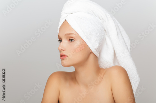Portrait of young beautiful woman with white towel on head without makeup thoughtfully looking aside with concealer cream on cheek over gray background isolated