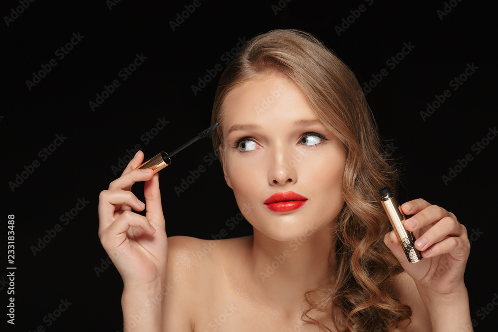 Portrait of young beautiful lady with wavy hair and red lips holding mascara in hand while dreamily looking aside over black background