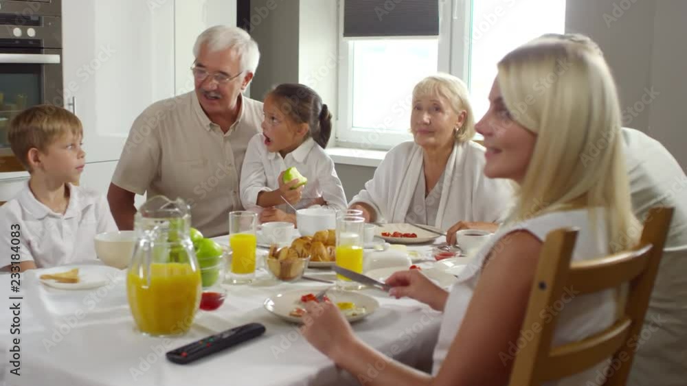 Beautiful family enjoying breakfast and watching TV in the morning: granddad bonding and talking with kids and mother and father smiling and looking at screen Beautiful family enjoying breakfast and watching TV in the morning: granddad bonding and talking with kids and mother and father smiling and looking at screen