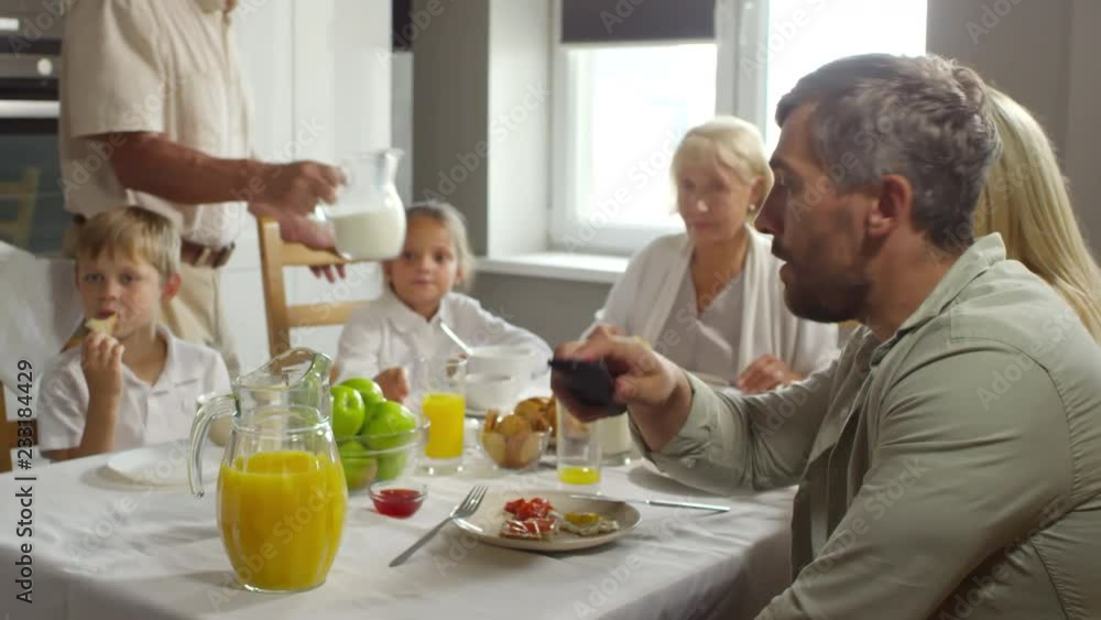 Dad turning on TV and watching program while having family breakfast with wife, little daughter and son and grandparents at home Dad turning on TV and watching program while having family breakfast with wife, little daughter and son and grandparents at home