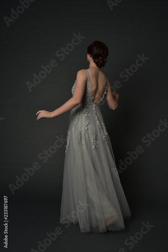 full length portrait of brunette girl wearing beautiful long gown, standing pose with background to the camera on grey studio background.