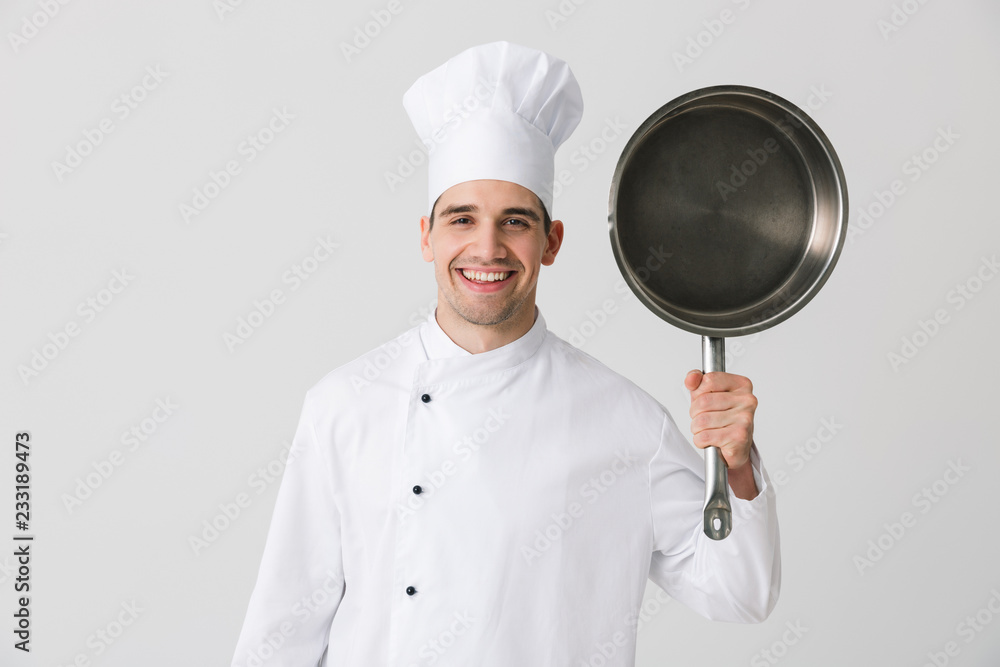 Emotional young man chef indoors isolated over white wall background holding frying pan. Emotional young man chef indoors isolated over white wall background holding frying pan.
