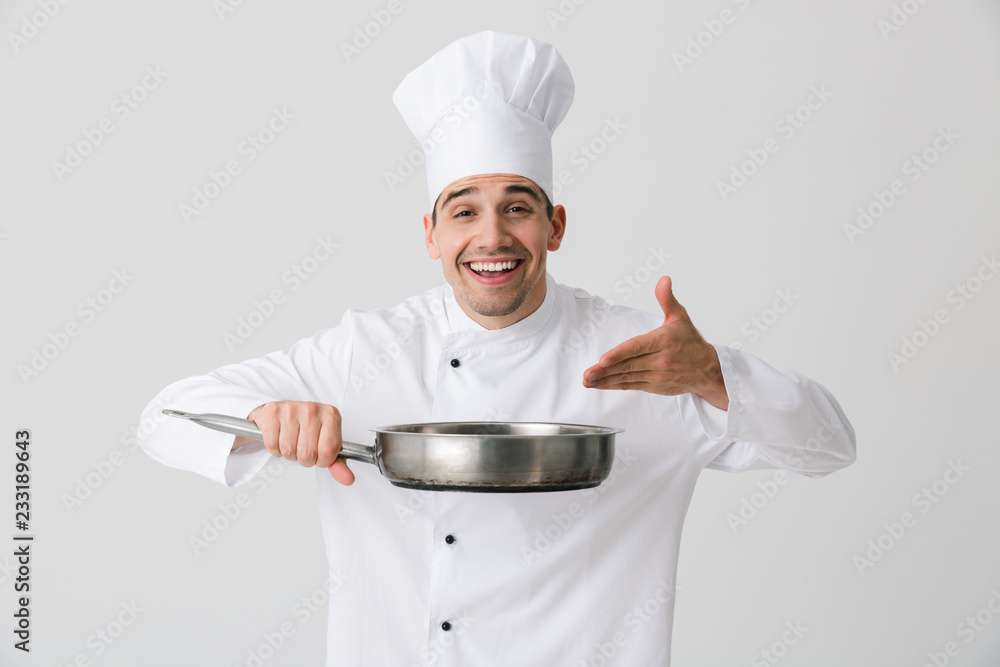Emotional young man chef indoors isolated over white wall background holding frying pan. Emotional young man chef indoors isolated over white wall background holding frying pan.