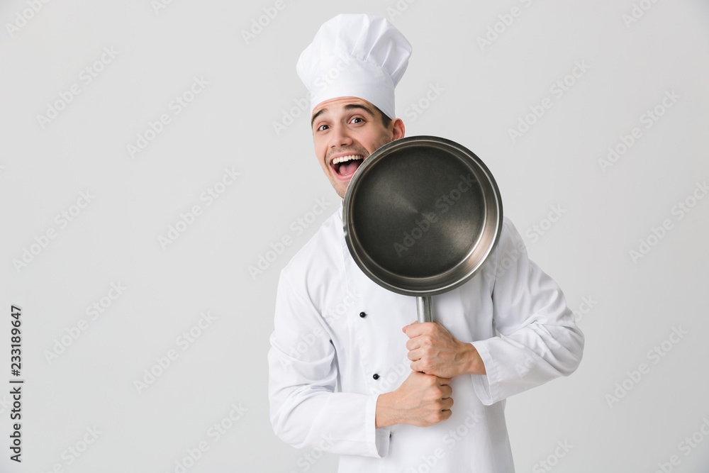 Emotional young man chef indoors isolated over white wall background holding frying pan. Emotional young man chef indoors isolated over white wall background holding frying pan.