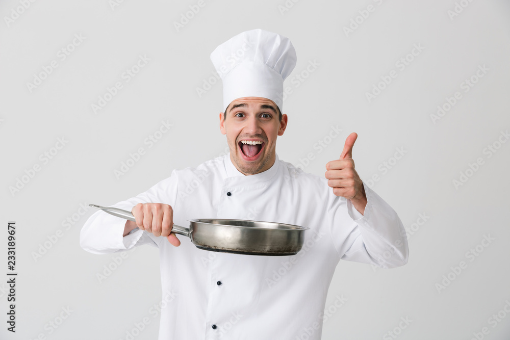 Emotional young man chef indoors isolated over white wall background holding frying pan. Emotional young man chef indoors isolated over white wall background holding frying pan.