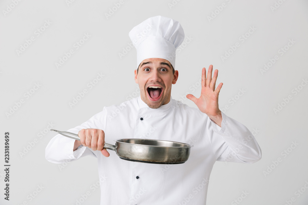 Emotional young man chef indoors isolated over white wall background holding frying pan. Emotional young man chef indoors isolated over white wall background holding frying pan.