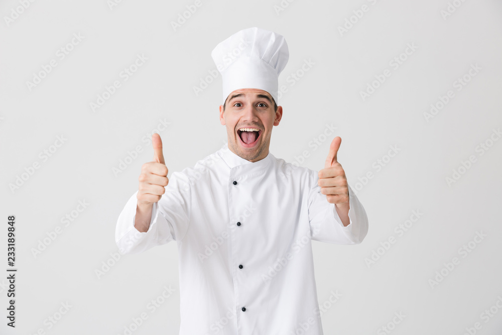 Excited young man chef indoors isolated over white wall background showing thumbs up gesture. Excited young man chef indoors isolated over white wall background showing thumbs up gesture.