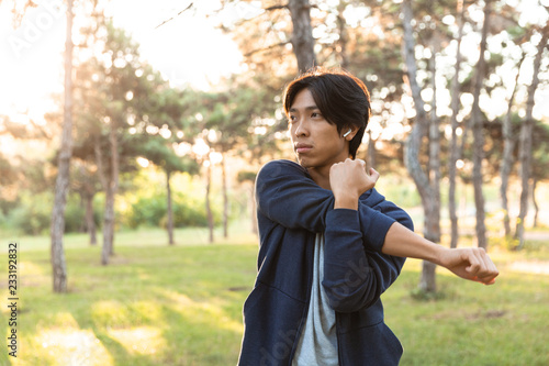 Image of sporty asian guy in casual wear stretching his body, while doing sports in green park