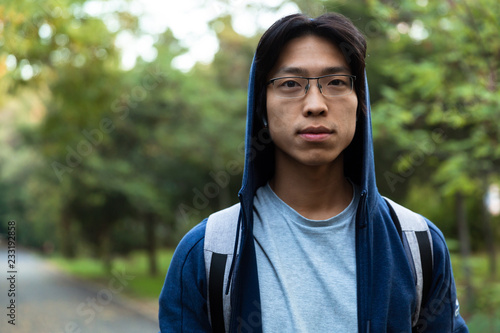 Image of young asian guy in casual wear and eyeglasses looking at you, while walking through green park