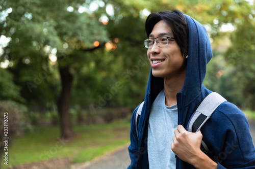 Image of positive asian man in casual wear and eyeglasses smiling, while walking through green park