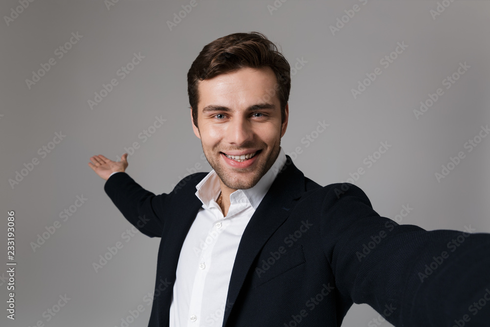 Image of optimistic businessman 30s in formal suit smiling on camera while taking selfie photo, isolated over gray background Image of optimistic businessman 30s in formal suit smiling on camera while taking selfie photo, isolated over gray background