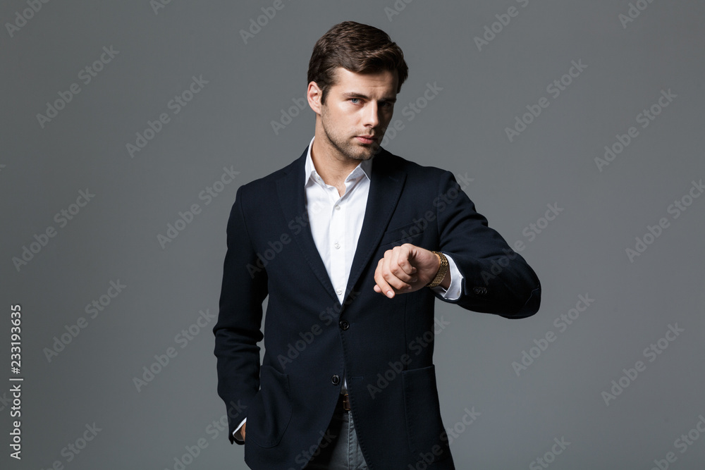 Image of young businessman 30s in black jacket looking at wristwatch, isolated over gray background Image of young businessman 30s in black jacket looking at wristwatch, isolated over gray background
