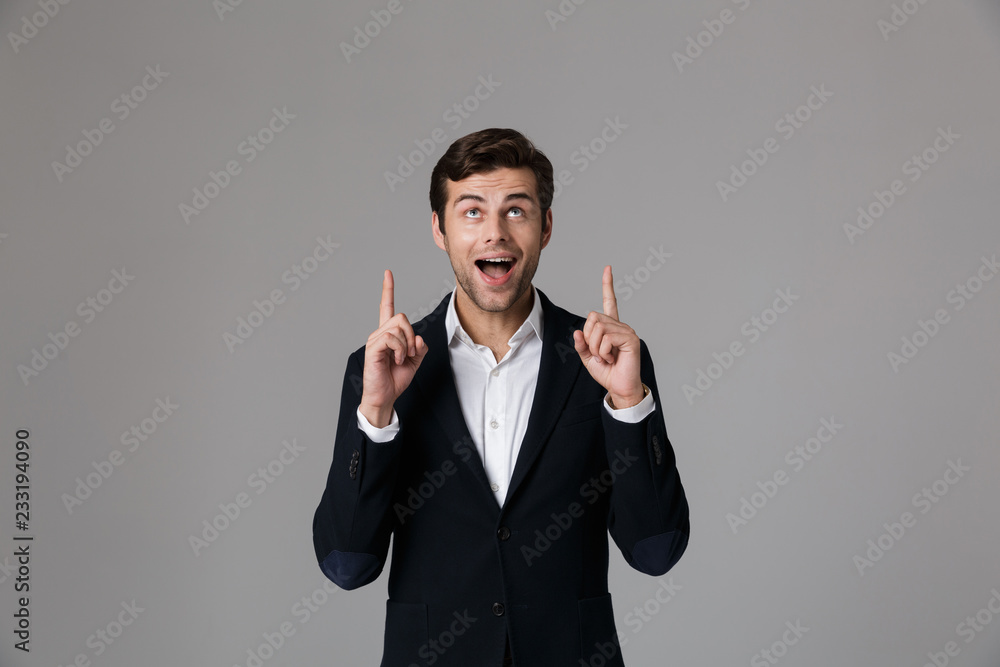 Image of joyful businessman 30s in formal suit pointing fingers upward, isolated over gray background Image of joyful businessman 30s in formal suit pointing fingers upward, isolated over gray background