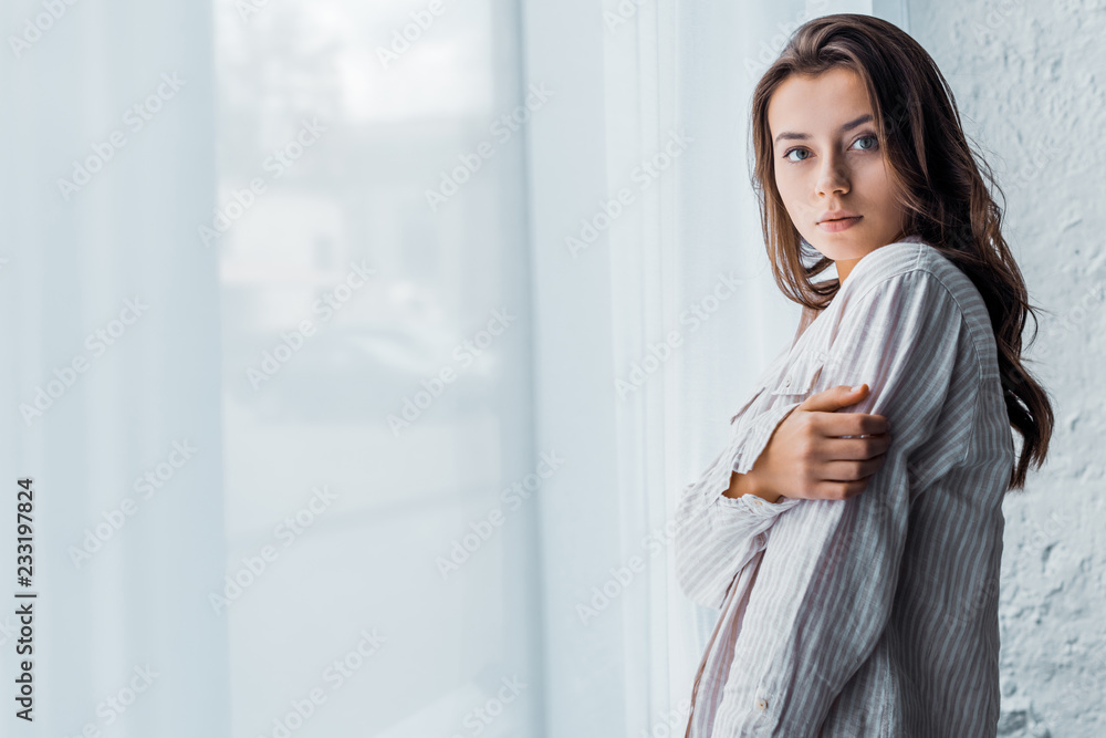 brunette tender girl in pajamas standing near window with white curtain