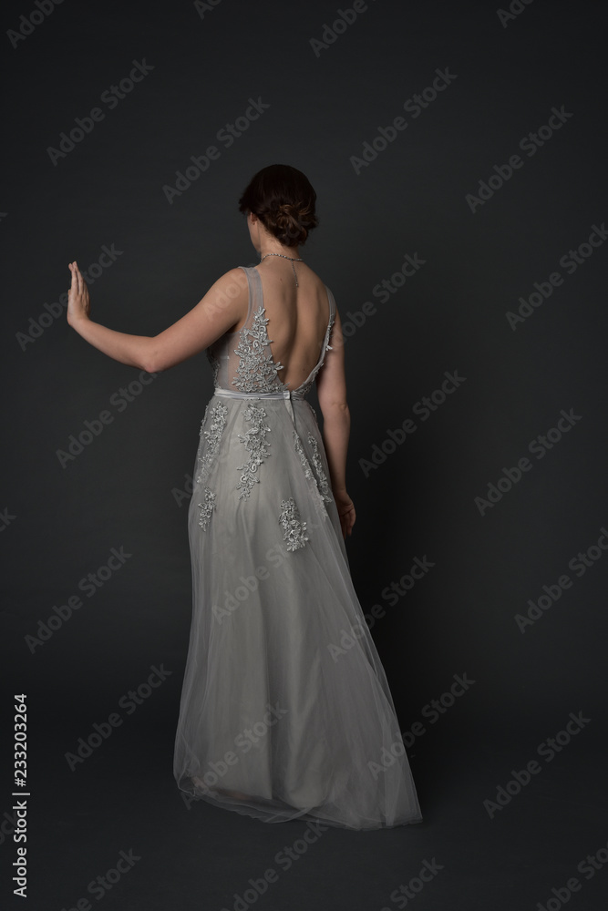 full length portrait of brunette girl wearing beautiful long gown, standing pose with background to the camera on grey studio background. full length portrait of brunette girl wearing beautiful long gown, standing pose with background to the camera on grey studio background.