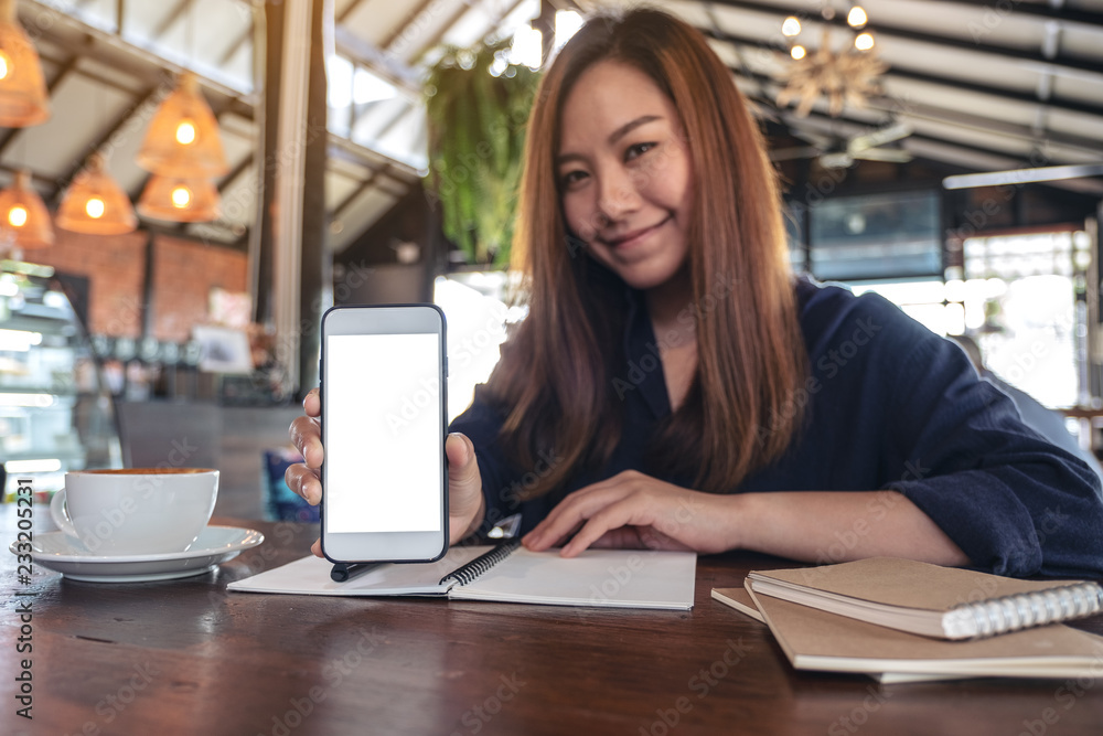 Mockup image of an asian woman holding and showing white mobile phone with blank desktop screen with coffee cups and notebooks on vintage wooden table in cafe Mockup image of an asian woman holding and showing white mobile phone with blank desktop screen with coffee cups and notebooks on vintage wooden table in cafe