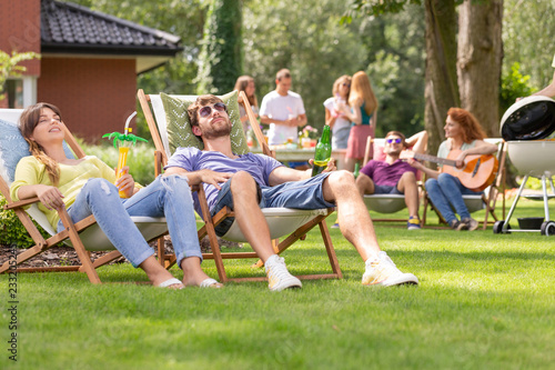 Man and woman relaxing on sunbeds during grill party with friends in the garden