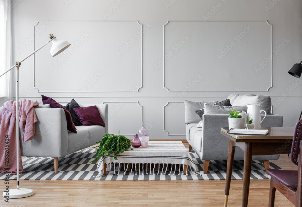 Obraz premium White lamp in stylish living room interior with two sofas with pillows and coffee table with green plant in pot, real photo with copy space on the empty wall