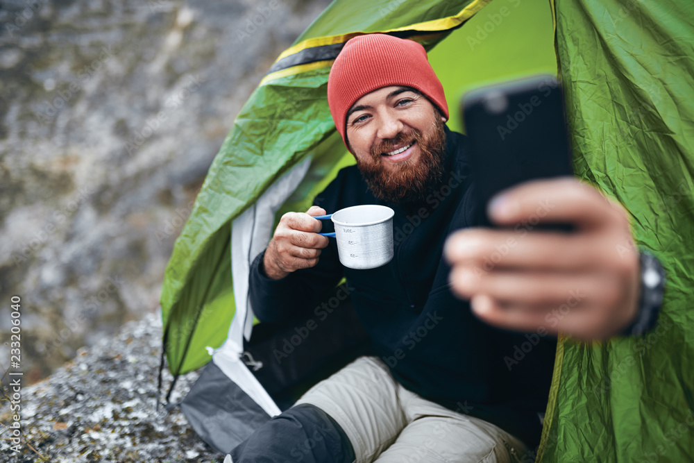 Young bearded man smiling, drinking tea and taking selfie in mountains ...