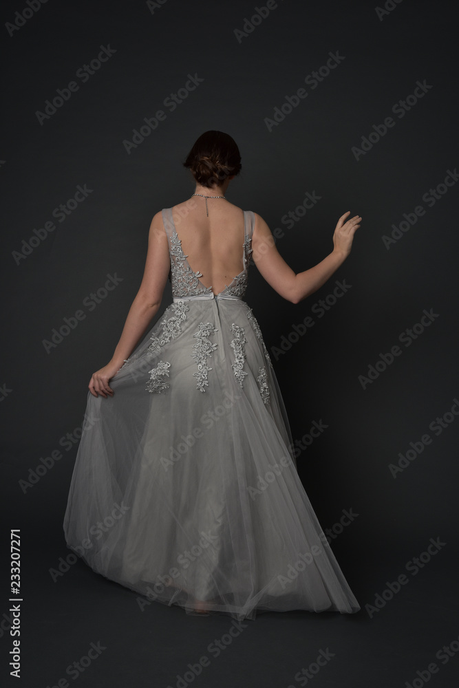 full length portrait of brunette girl wearing beautiful long gown, standing pose with background to the camera on grey studio background. full length portrait of brunette girl wearing beautiful long gown, standing pose with background to the camera on grey studio background.
