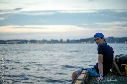 Portrait Asian man sitting on the port and looking out to the sea with the twilight sky of sunset and bokeh lights of the city in background