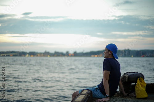 Portrait Asian man sitting on the port and looking out to the sea with the twilight sky of sunset and bokeh lights of the city in background