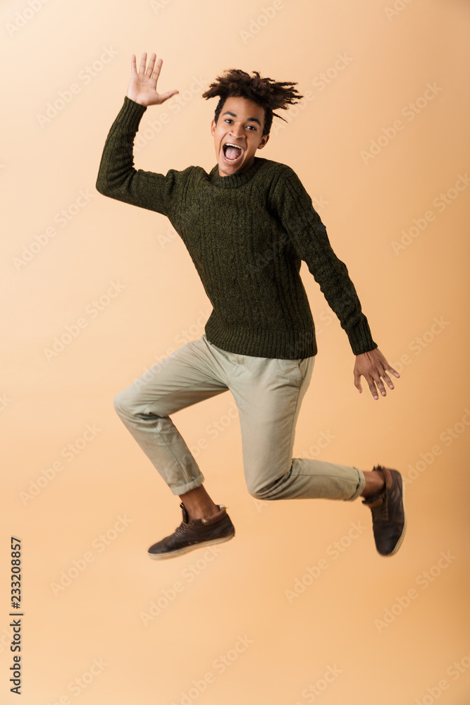 Full length photo of amusing african american guy wearing sweater laughing and jumping, isolated over beige background Full length photo of amusing african american guy wearing sweater laughing and jumping, isolated over beige background