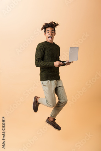 Full length photo of pleased african american guy wearing sweater walking with laptop, isolated over beige background