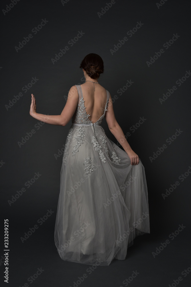 Fototapeta premium full length portrait of brunette girl wearing beautiful long gown, standing pose with background to the camera on grey studio background.