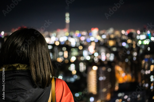 Young asian woman with short black hair is standing on a observation deck and looks at the night Tokyo (blurry background)