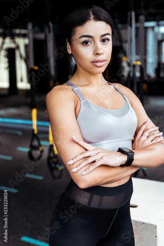 Beautiful sportive girl with crossed arms llooking at camera while standing in fitness gym