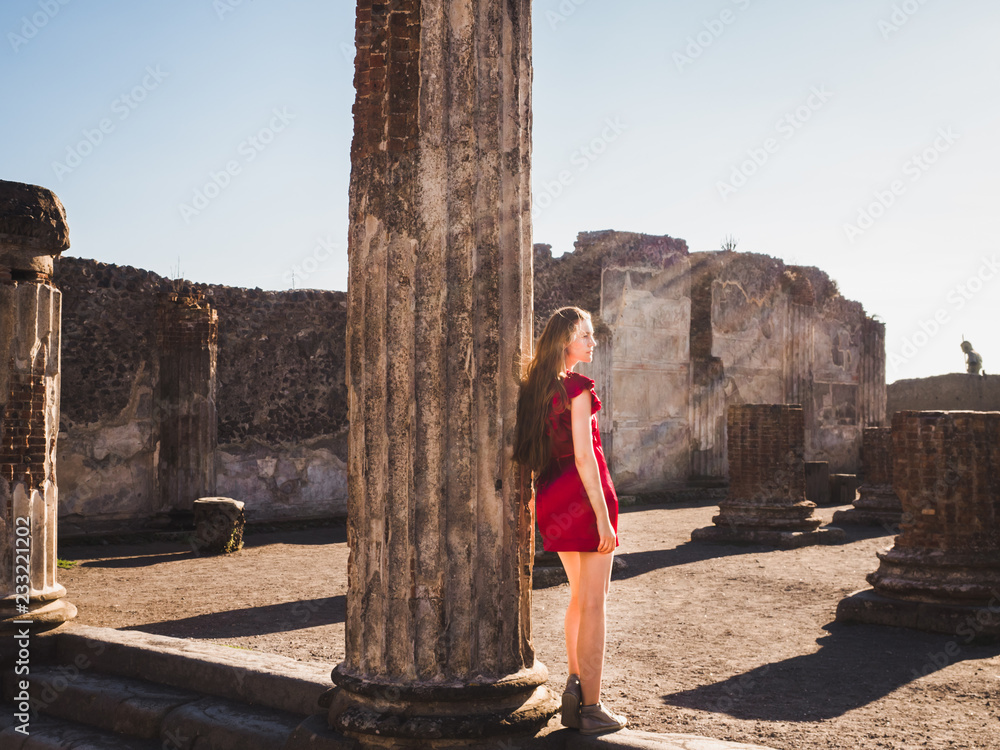 Stylish woman in a red dress looking into the distance against the backdrop of ancient ruins and the sun's rays of the evening sun. Pompeii. Concept of travel and leisure