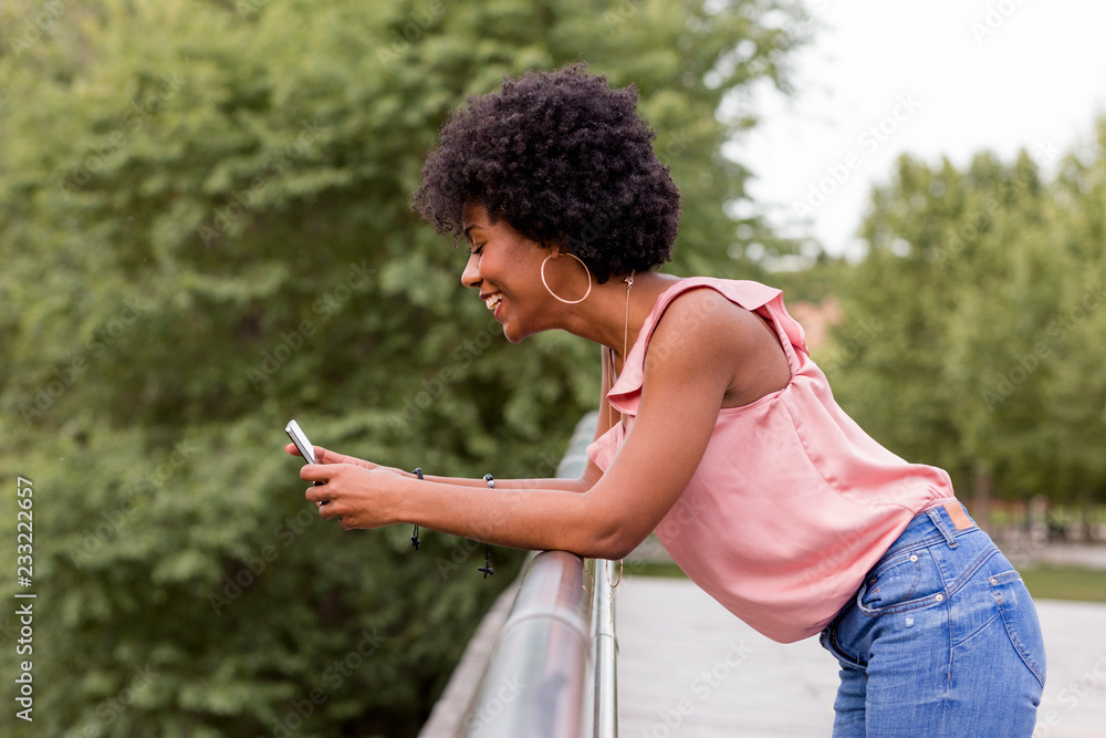 Happy young beautiful afro american woman smiling and using mobile phone. Green background. Spring or summer season. Casual clothing outdoors Happy young beautiful afro american woman smiling and using mobile phone. Green background. Spring or summer season. Casual clothing outdoors