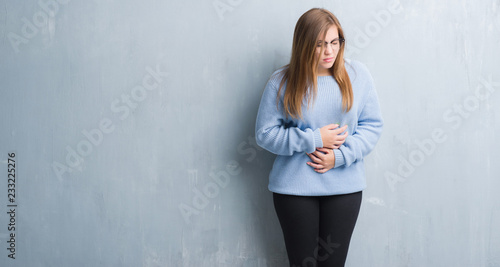 Young adult woman over grey grunge wall wearing glasses with hand on stomach because nausea, painful disease feeling unwell. Ache concept.