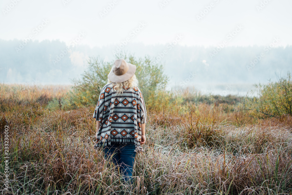 Woman in wide-brimmed felt hat and authentic poncho standing in high grass at foggy morning