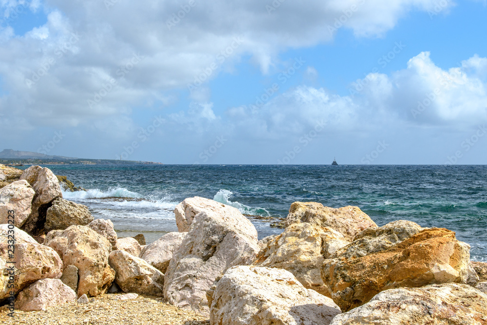 Seascape with huge rocks in the foreground, surf waves, ship silhouette on the horizon and blue sky with clouds. Seascape with huge rocks in the foreground, surf waves, ship silhouette on the horizon and blue sky with clouds.