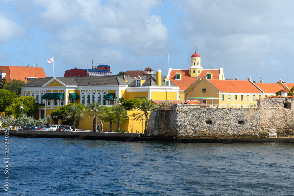 View from the sea to the embankment of the city with yellow houses under the red roof, palm trees and a stone wall of the ancient fortress
