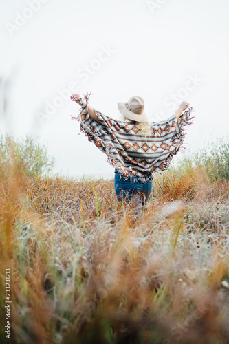 Woman in wide-brimmed felt hat and authentic poncho standing in high grass at foggy morning