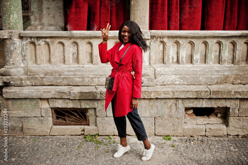 Stylish african american woman in red coat posed against old columns and show two fingers sign.